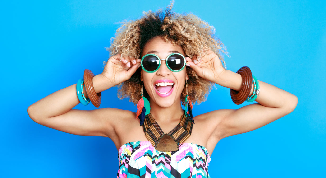 Curly-haired woman smiles against a blue wall while wearing sunglasses to welcome new patients to Megan Jones, DDS