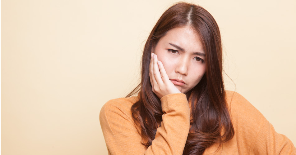 Brunette woman in a yellow shirt cringes in pain and touches her cheek due to a toothache