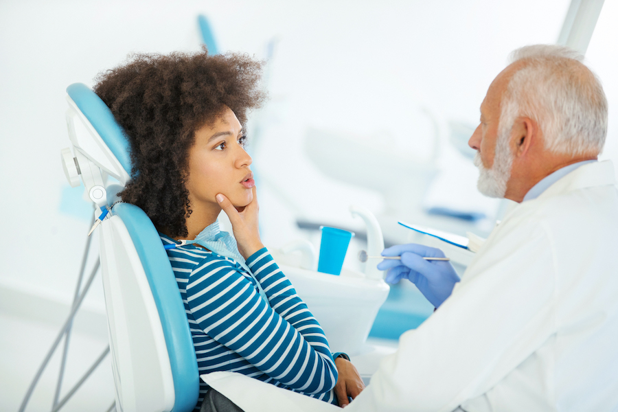 Black woman in a dental chair touches her jaw and consults with her white older male dentist