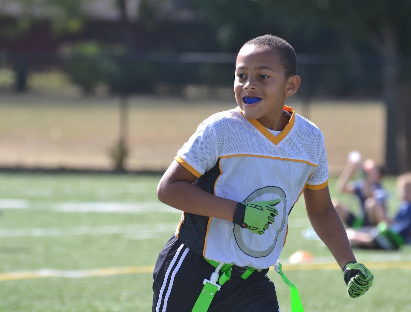 A young Black boy wears a mouthguard while playing flag football outside on a green field