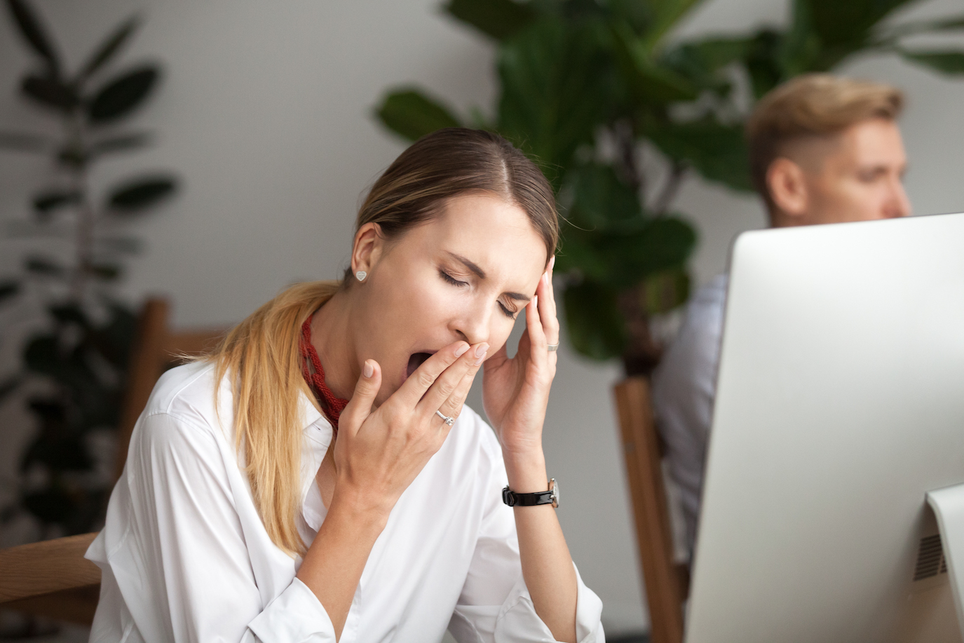 Blonde woman in a white shirts yawns at her desk during the day due to chronic fatigue