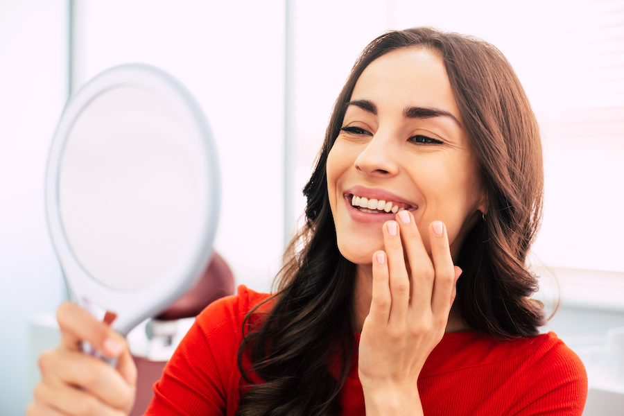 A brunette woman in a red blouse smiles at herself in a small hand mirror