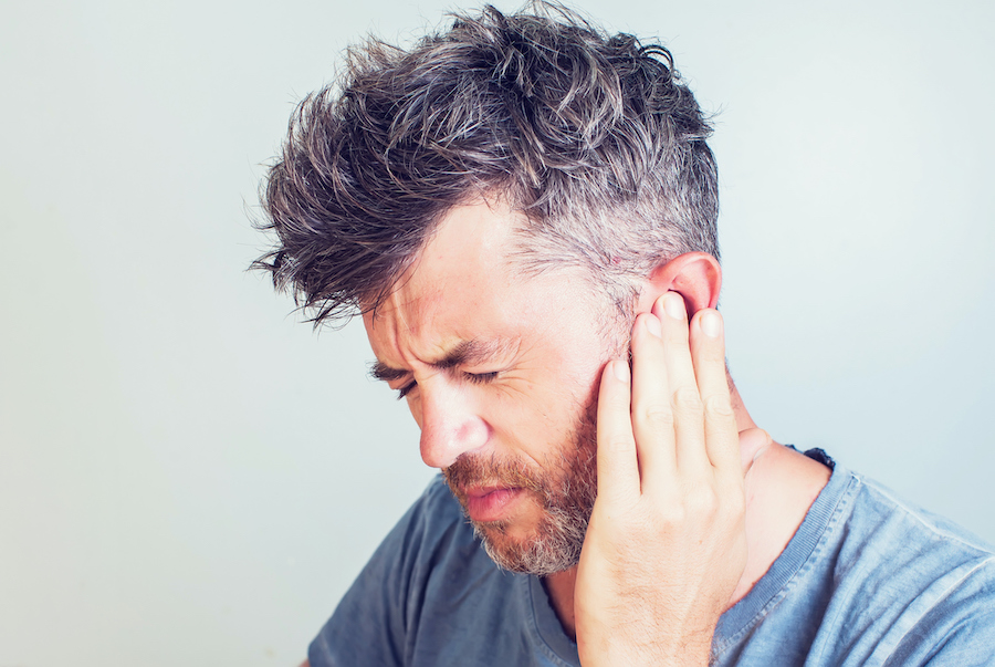 Middle-aged man in a gray shirt cringes in pain and touches his ear because of an earache