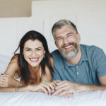 Brunette woman and man with salt and pepper hair smile with teeth improved with dental restorations